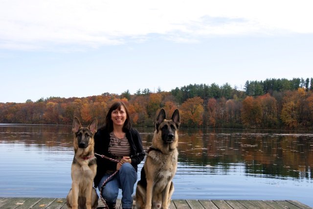 Person kneeling on a wooden dock with two German Shepherds beside a calm lake surrounded by autumn trees.